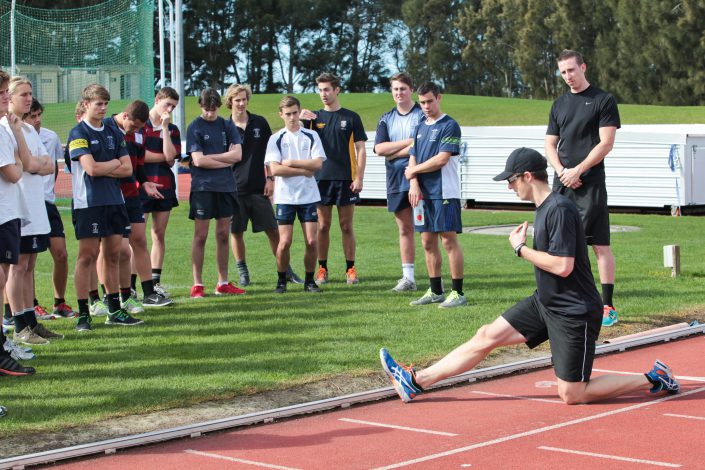 Dr Craig Harrison in front of a group of male athletes demonstrating a stretch.