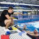 Male coach Jacob Garrod pool side coaching with flippers and flotation devices around him. Two middle aged males with googles on are holding onto the side of the pool listening to Jacob's instruction